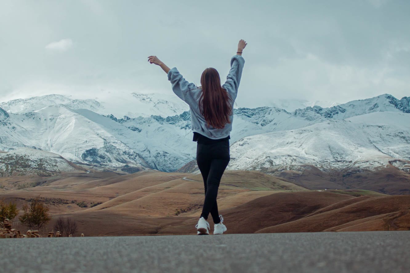 A woman on a country road with her arms raised, facing snow-covered mountains.