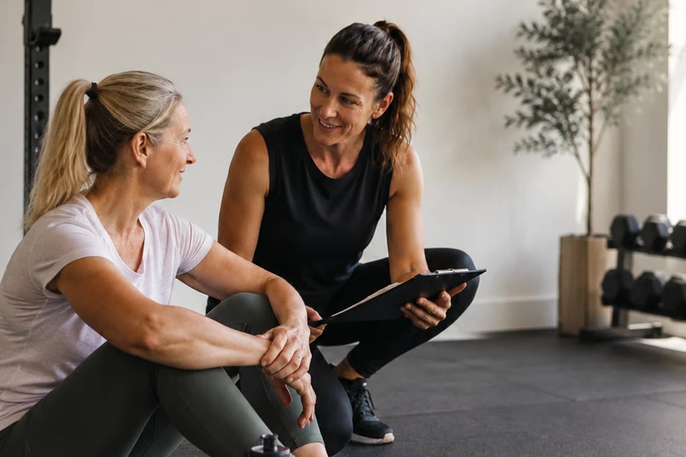 An exercise professional with a clipboard, sitting and talking with a client on a gym floor.